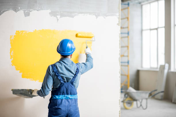 Workman in uniform painting wall with yellow paint at the construction site indoors